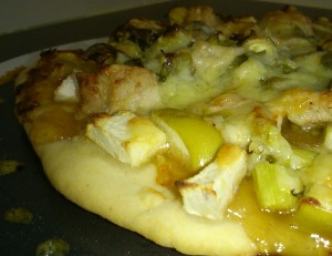 A pizza. The base is topped with a clear brown sauce, then apples and pieces of white meat, and finally cheese.
